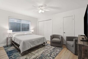 Una camera da letto con un letto, due poltrone, una TV, una ventola a soffitto e una finestra con persiane.