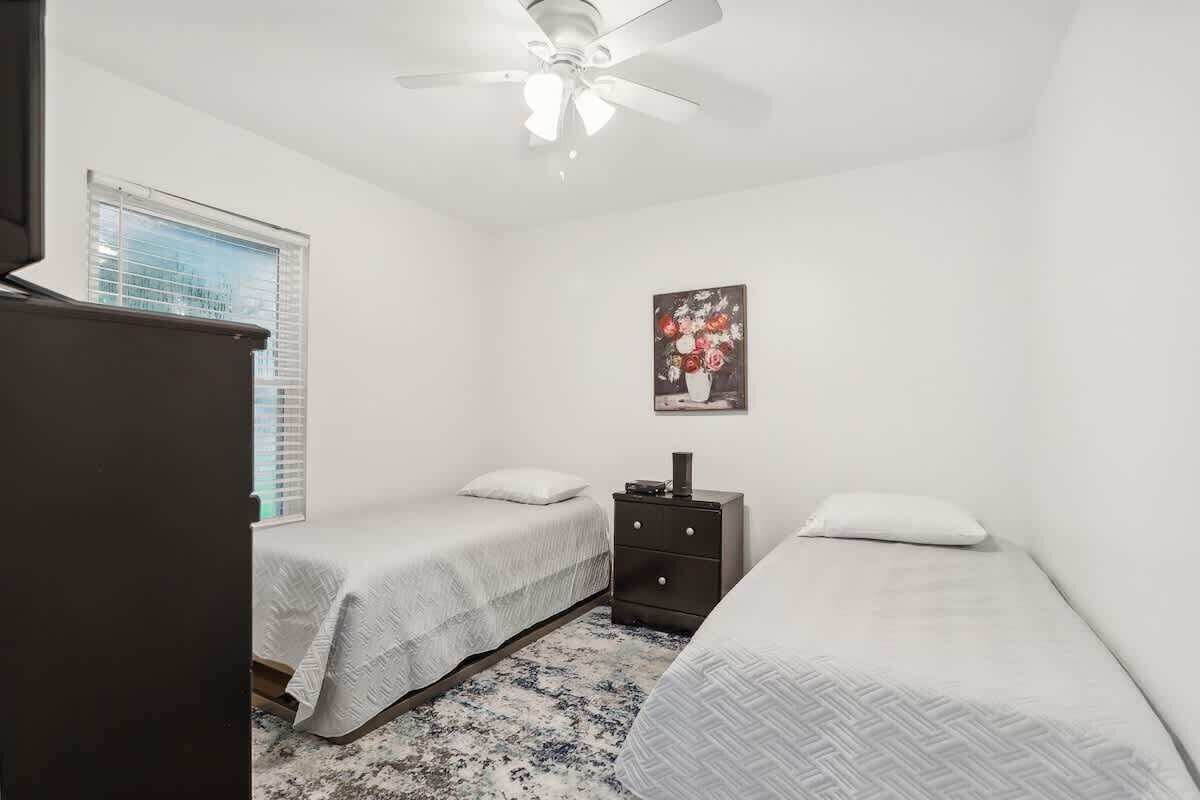 A bedroom with two single beds, a ceiling fan, a dresser, and a window with blinds.