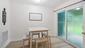 Ein Zimmer mit einem weißen Tisch und Bänken, einem gerahmten Zitat an der Wand und einer Glasschiebetür zu einem grünen Außenbereich.