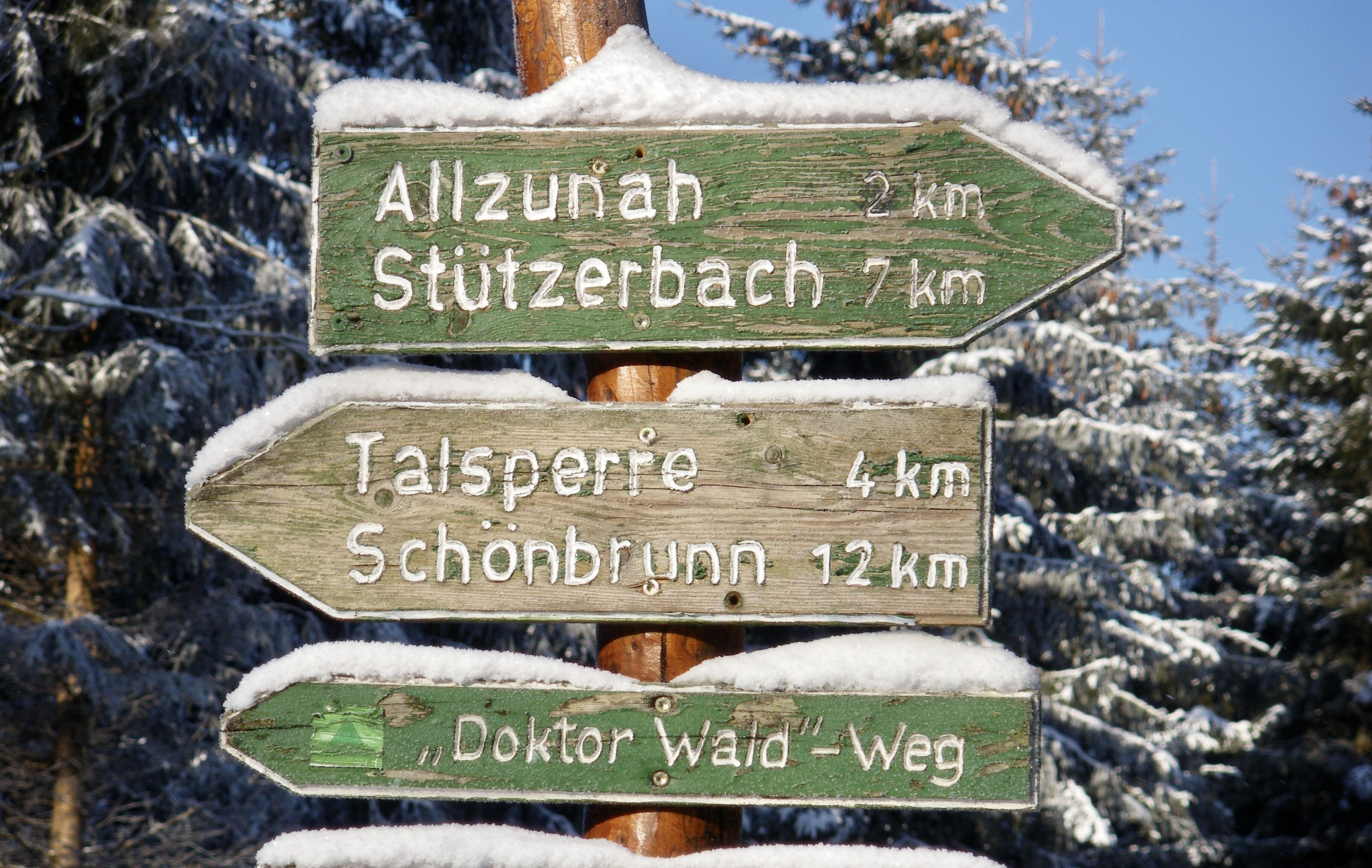 A wooden directional signpost with distance markers for Allzunah, Stutzerbach, Talsperre, Schonbrunn, and Doktor Wald-Weg.