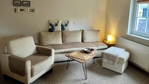 Una sala de estar con sofá, sillón y reposapiés, una mesita de madera y un ventanal con vistas.