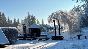 Un espacio al aire libre cubierto de nieve, con mesas de picnic, un pequeño refugio y árboles cargados de nieve.