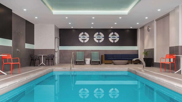 A modern indoor pool area with a clear pool, seating by the pool, and a wall with circular logos. - Tru By Hilton Bracebridge, On (Bracebridge)