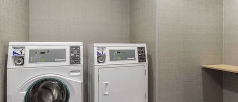 Two front-loading washing machines in a laundry room with grey walls and a wooden shelf.
