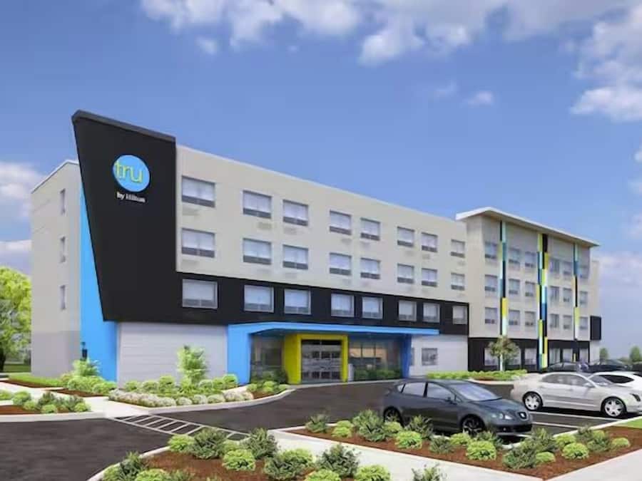 A modern hotel building with a prominent logo on the side, multiple floors, and a parking lot with several cars.