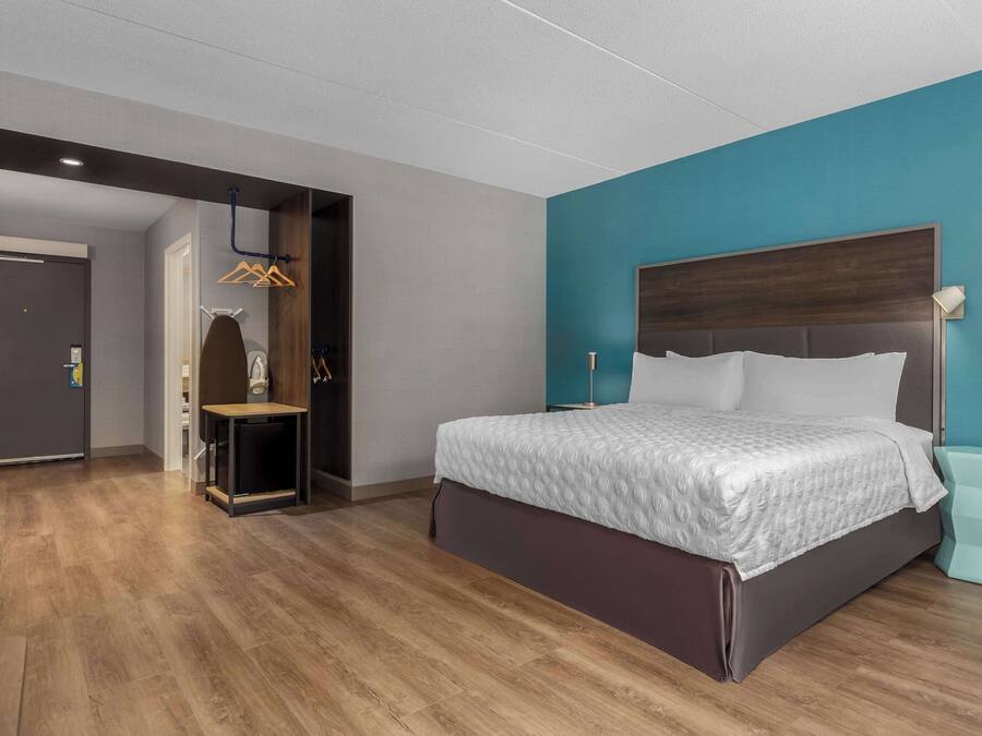 A modern hotel room with a large bed, a wooden floor, a teal accent wall, and a wardrobe with hanging clothes.