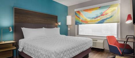 A modern hotel room with a large bed, a desk, a chair, and a colorful abstract painting.