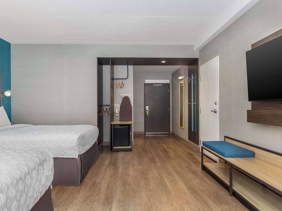 A modern hotel room with a bed, a bench, a TV, and a door leading to a bathroom.