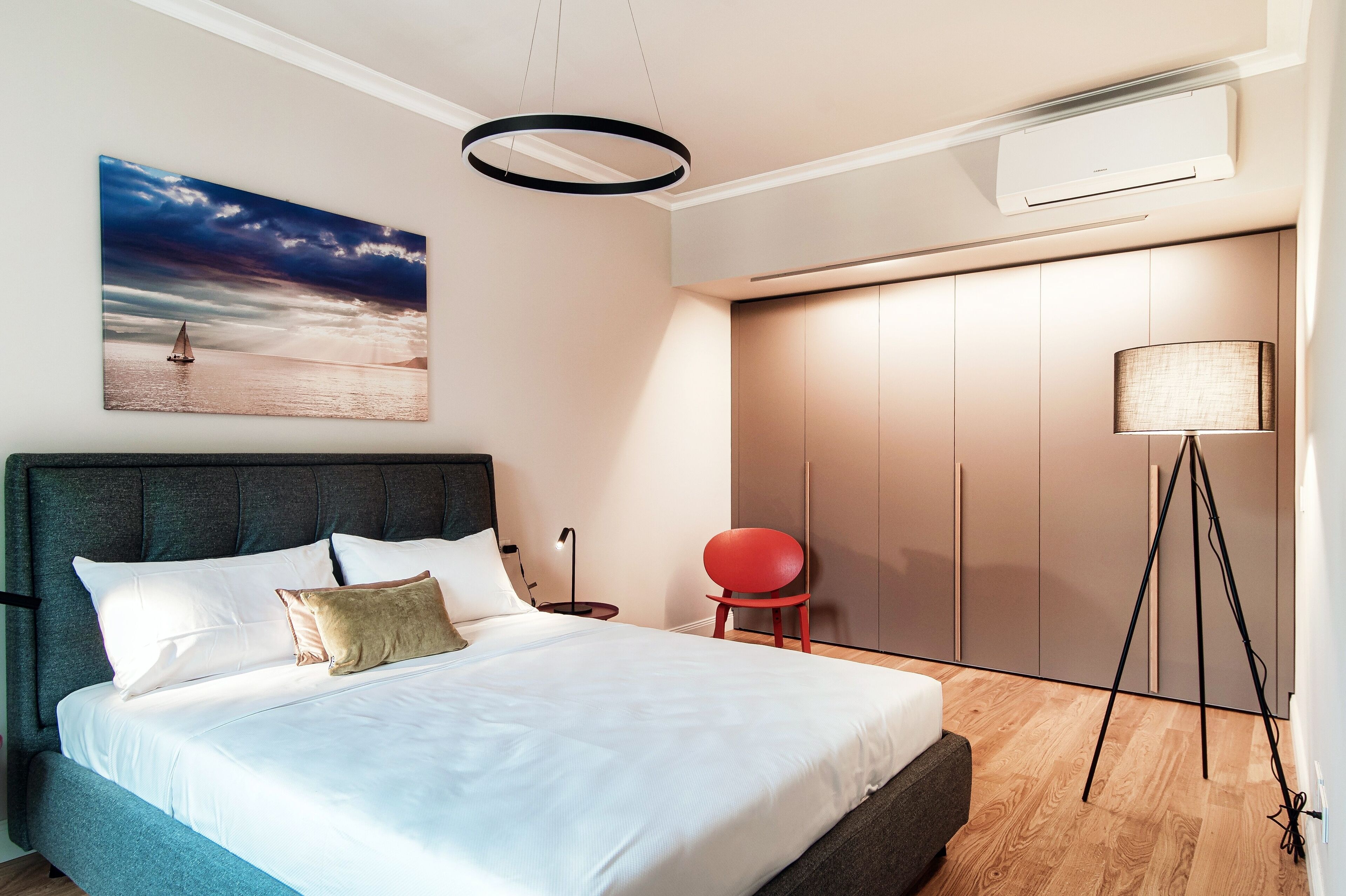 A modern bedroom with a large bed, a wall-mounted air conditioner, a floor lamp, a red chair, and a painting of a sailboat.