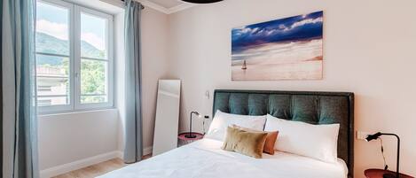 A neatly made bed with white linens and a green headboard, a bedside table with a lamp, and a painting of a sunset over water on the wall.