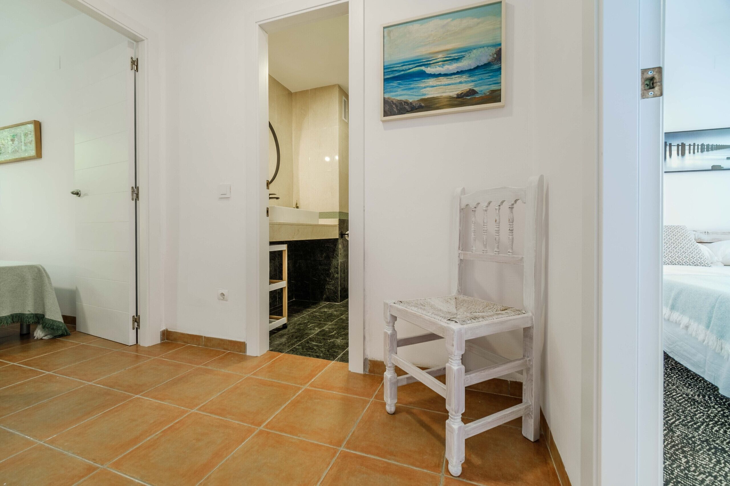 Quarto com uma cadeira branca, um quadro de mar e um banheiro visível por uma porta aberta.