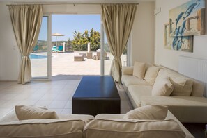 A living room with a sofa, a coffee table, and a view of an outdoor pool area. - Villa Marietta by Estia (Hersonissos)