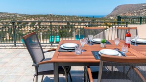 A wooden table set for four with metal chairs, a bottle of red wine, and a view of the sea and hills. - Villa Marietta by Estia (Hersonissos)