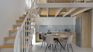 A modern kitchen with marble countertops, a dining area with a round table and chairs, and a wooden staircase with white railings. - Villa Chara by Estia (Mylopotamos)