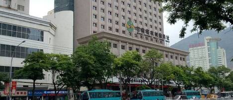 Un edificio de varios pisos con fachada plana, rodeado de árboles y una calle con autobuses estacionados.