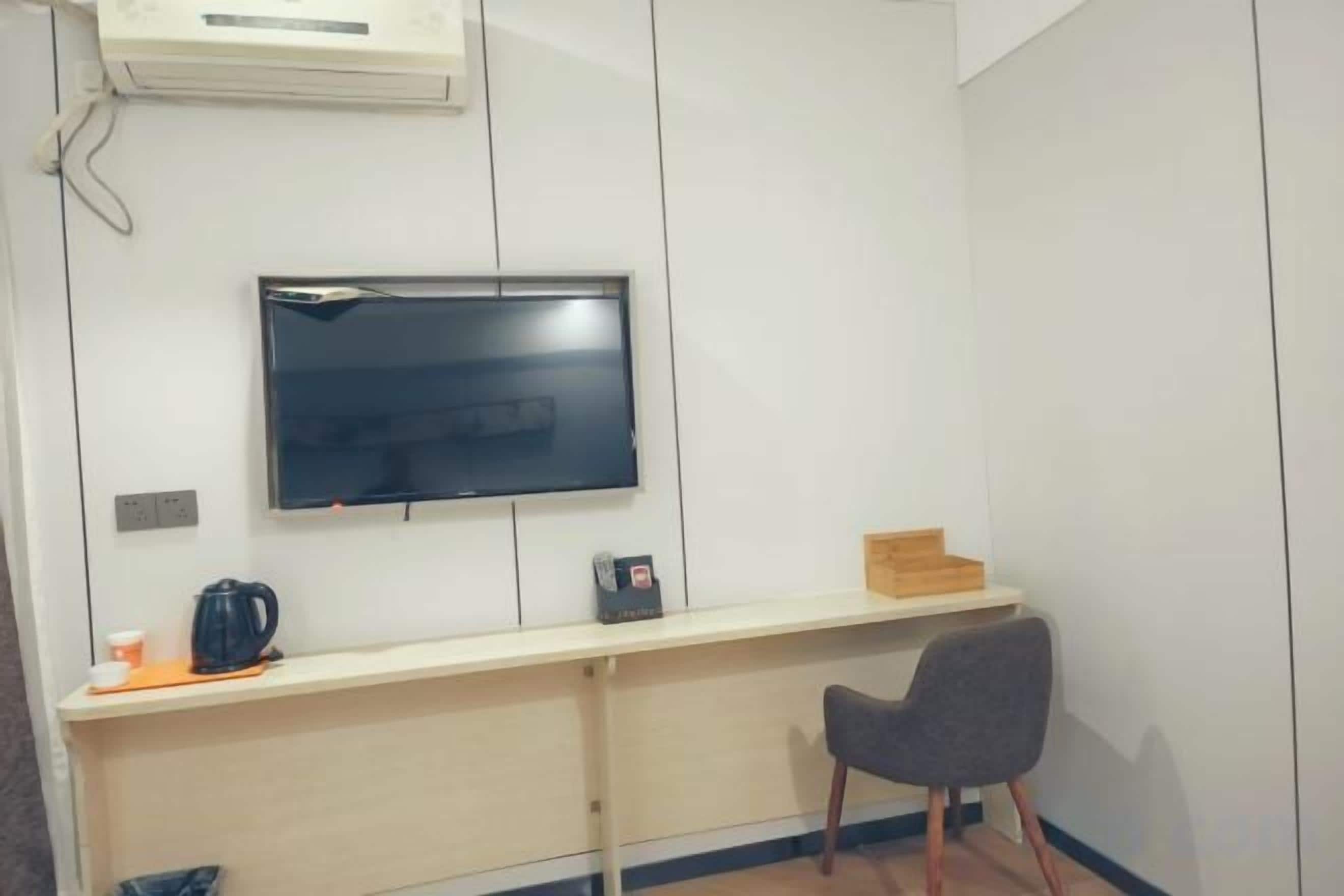 A room with a flat-screen TV mounted on the wall, a wooden desk, a chair, and a small table with a kettle and cup.