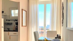 Una sala de estar con un sofá gris, un comedor con mesa y sillas, una cocina con armarios blancos y vistas a través de amplios ventanales.