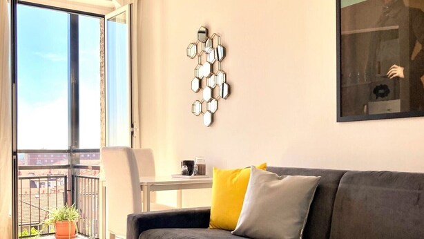 Una sala de estar con un sofá gris, una mesa de centro y una planta en maceta. Se ve un ventanal con cortinas y un balcón al exterior.