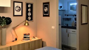 Un dormitorio con cama, mesita de noche con libros, lámpara y decoración en la pared. Se ve una cocina desde la habitación.