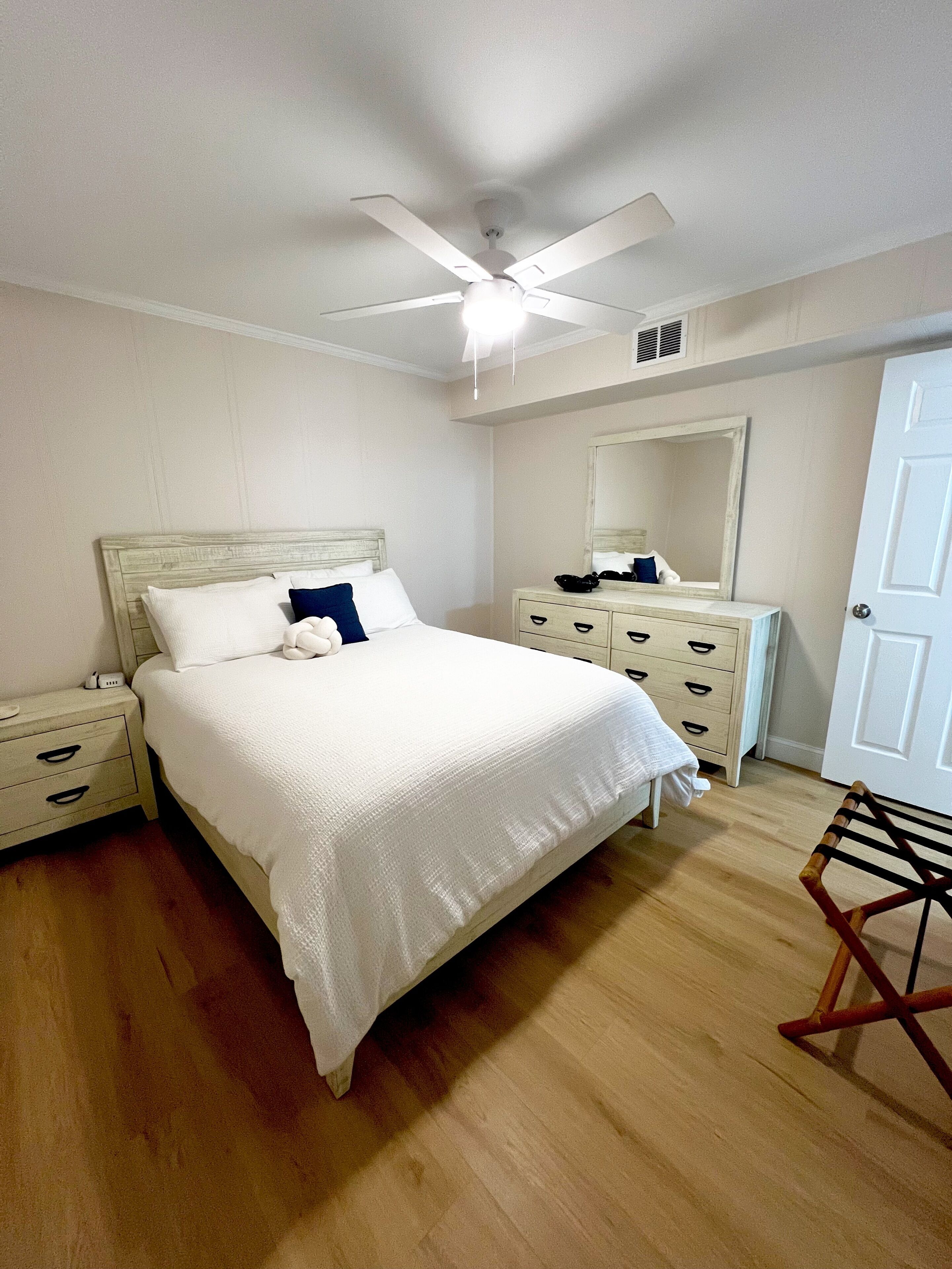 A bedroom with a bed, a nightstand, a dresser, a mirror, a ceiling fan, and a door.