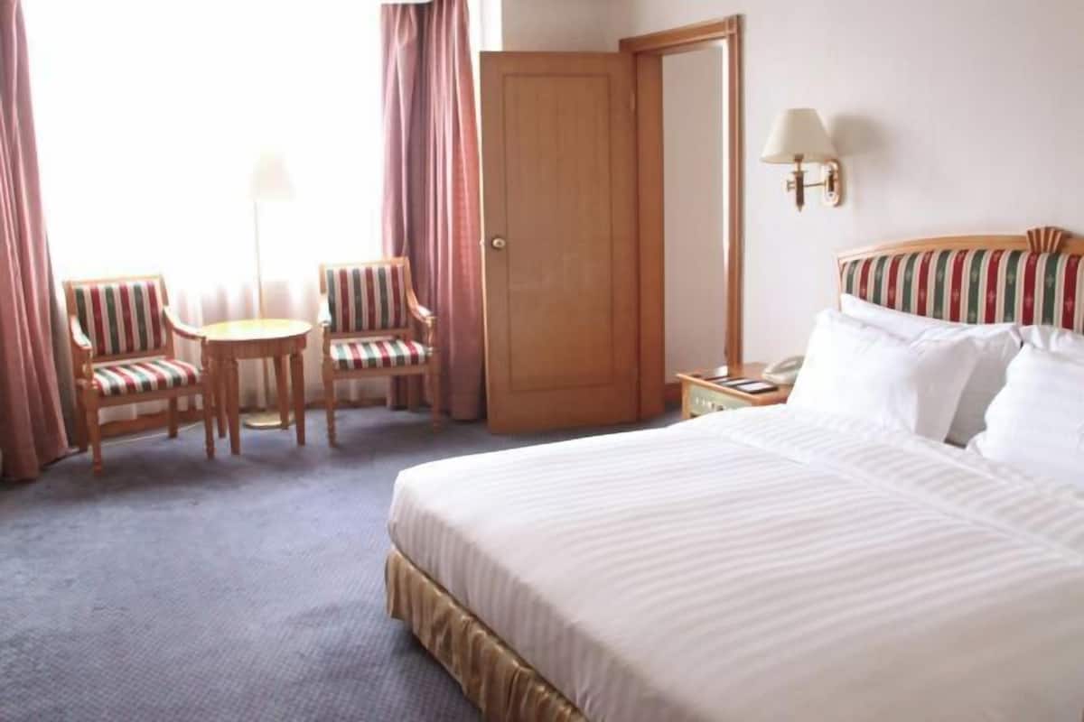 A hotel room with a bed, two striped chairs, a small table, a nightstand, a lamp, and a door.