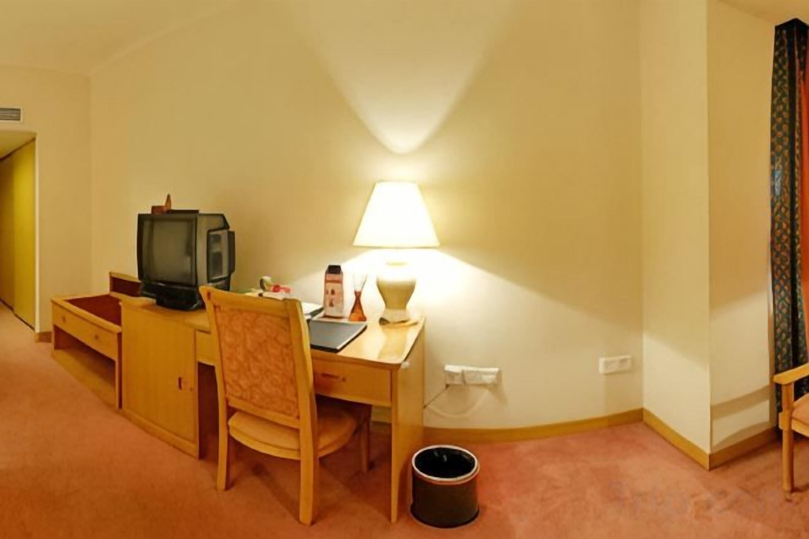 Una habitación pequeña con escritorio, silla, televisor, lámpara y un cubo de basura.