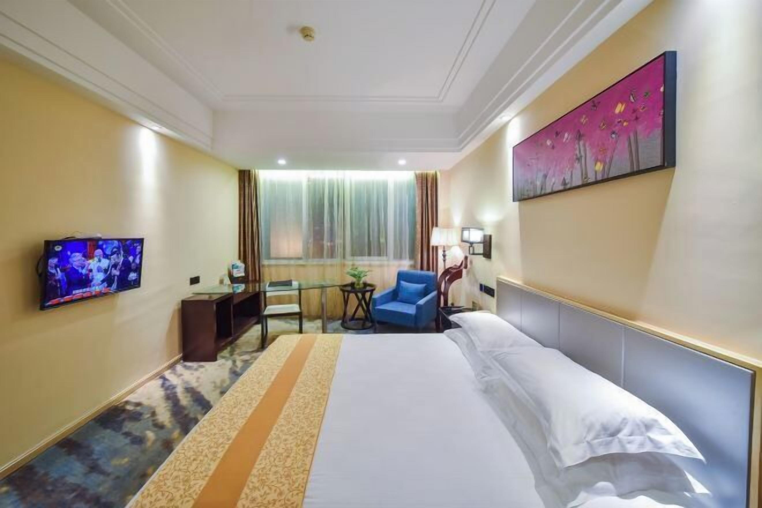 A hotel room with a bed, a TV, a desk, a chair, and a painting on the wall.