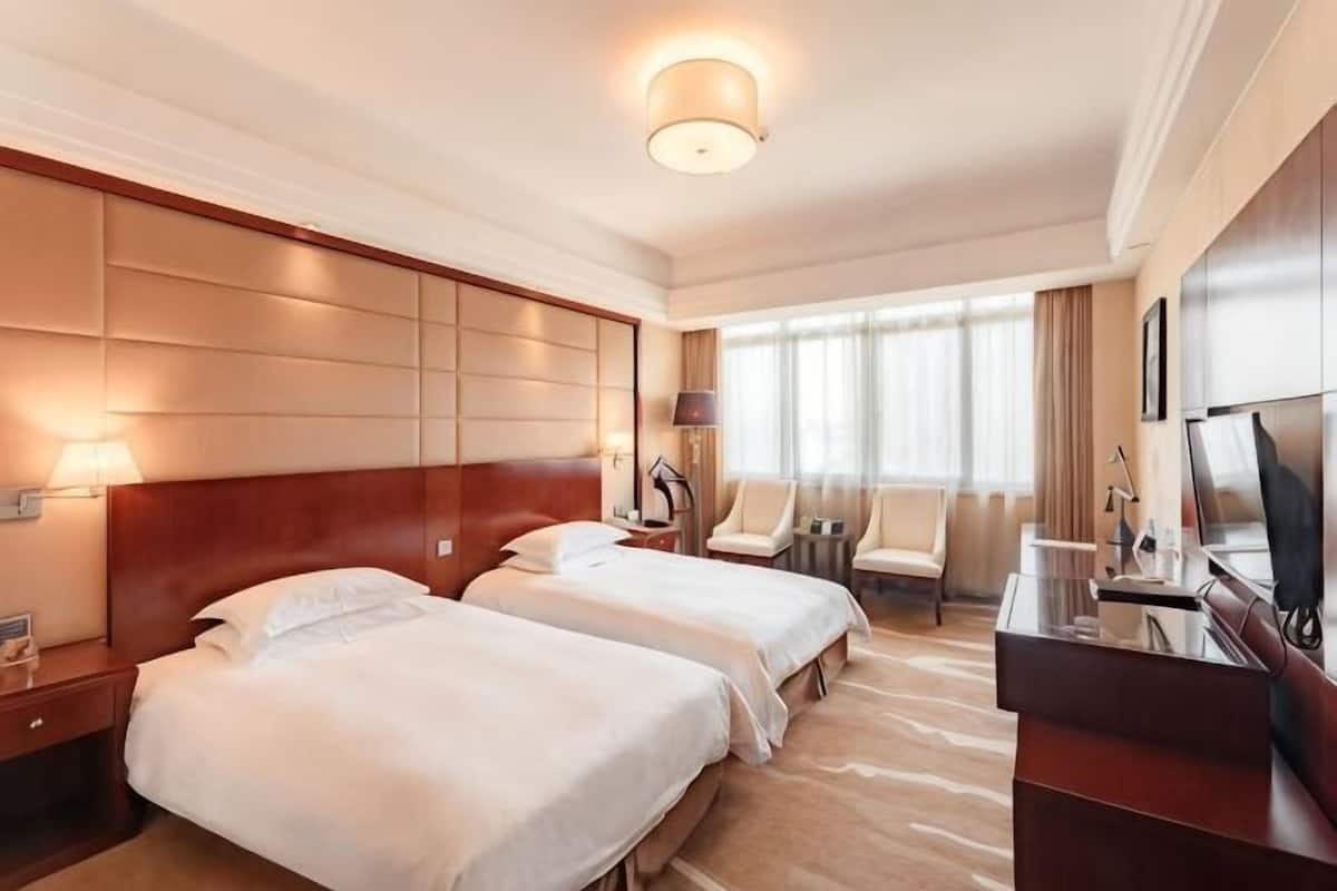 A hotel room with two beds, a desk, a TV, and a large window with curtains.