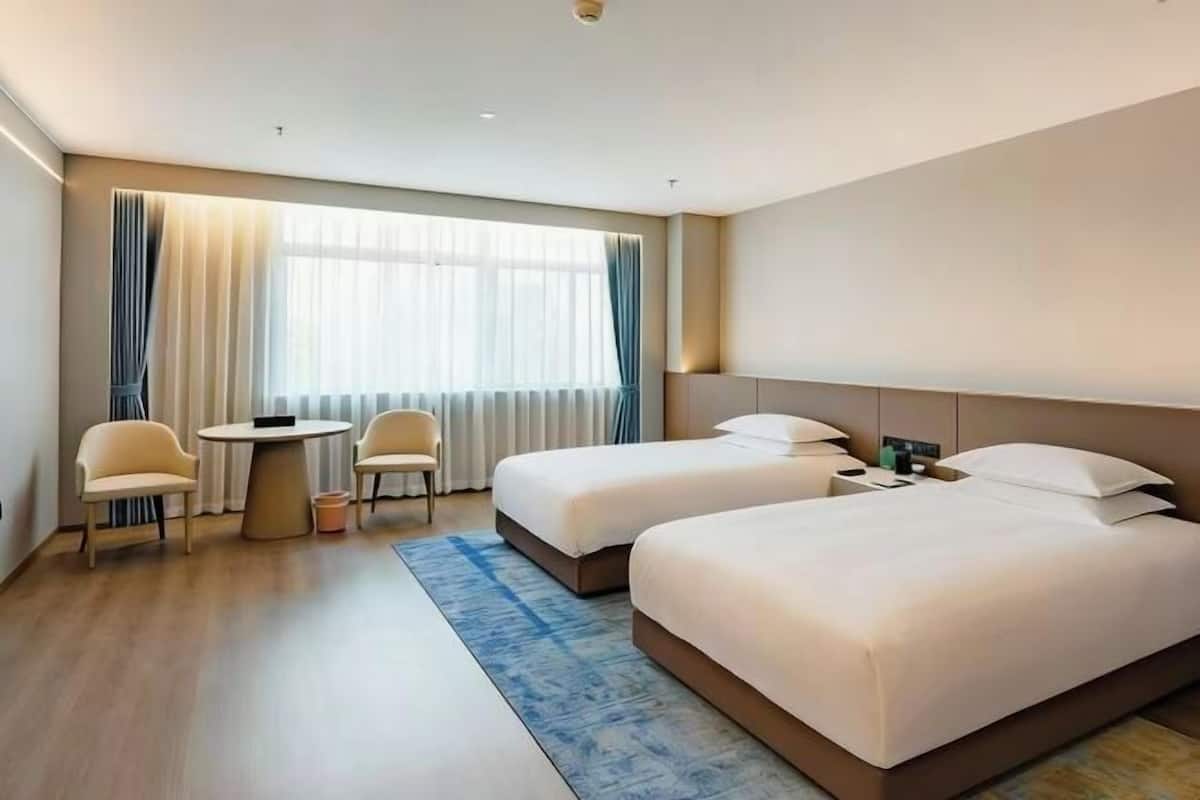 A hotel room with two beds, a small table, two chairs, a window with curtains, and a blue rug.