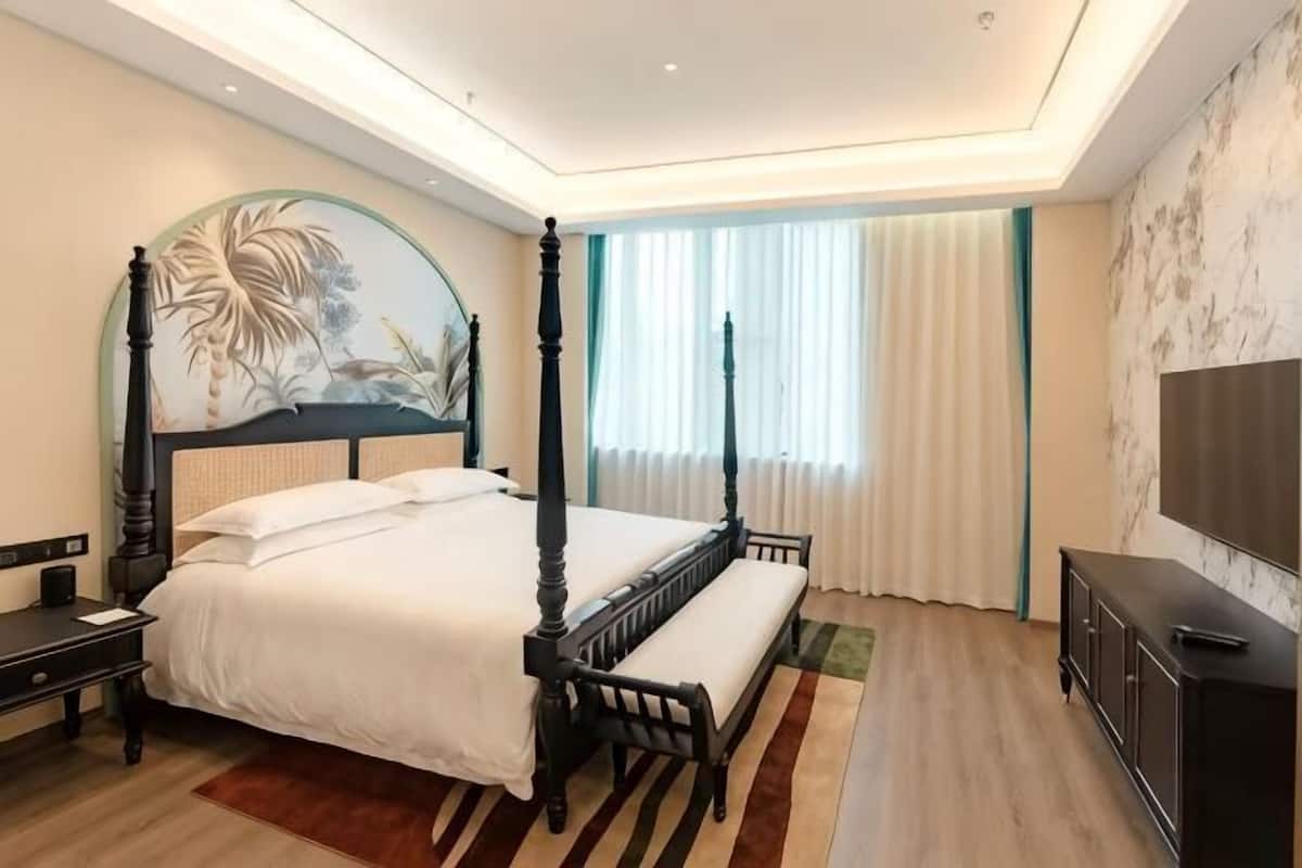 A four-poster bed with a headboard, a bedside table, a bench, a TV, and a large window with curtains.