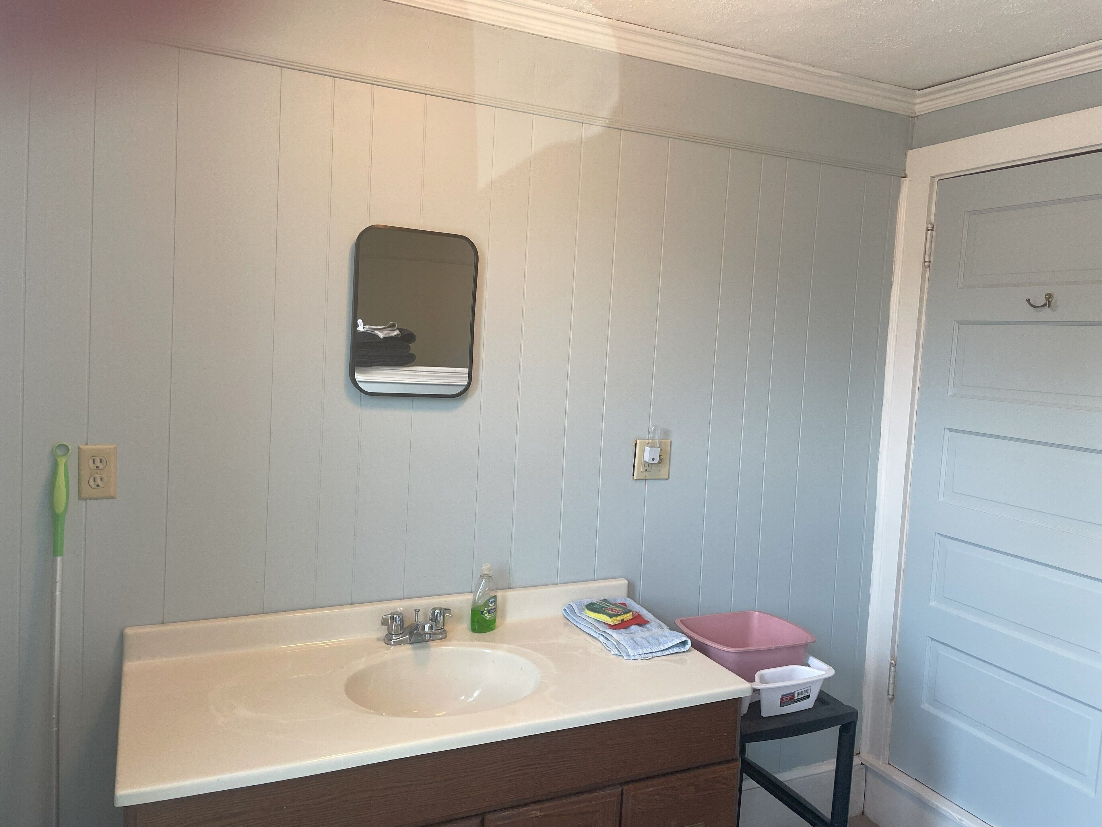 Un baño con un lavamanos blanco, un espejo y un taburete pequeño con un recipiente rosa.