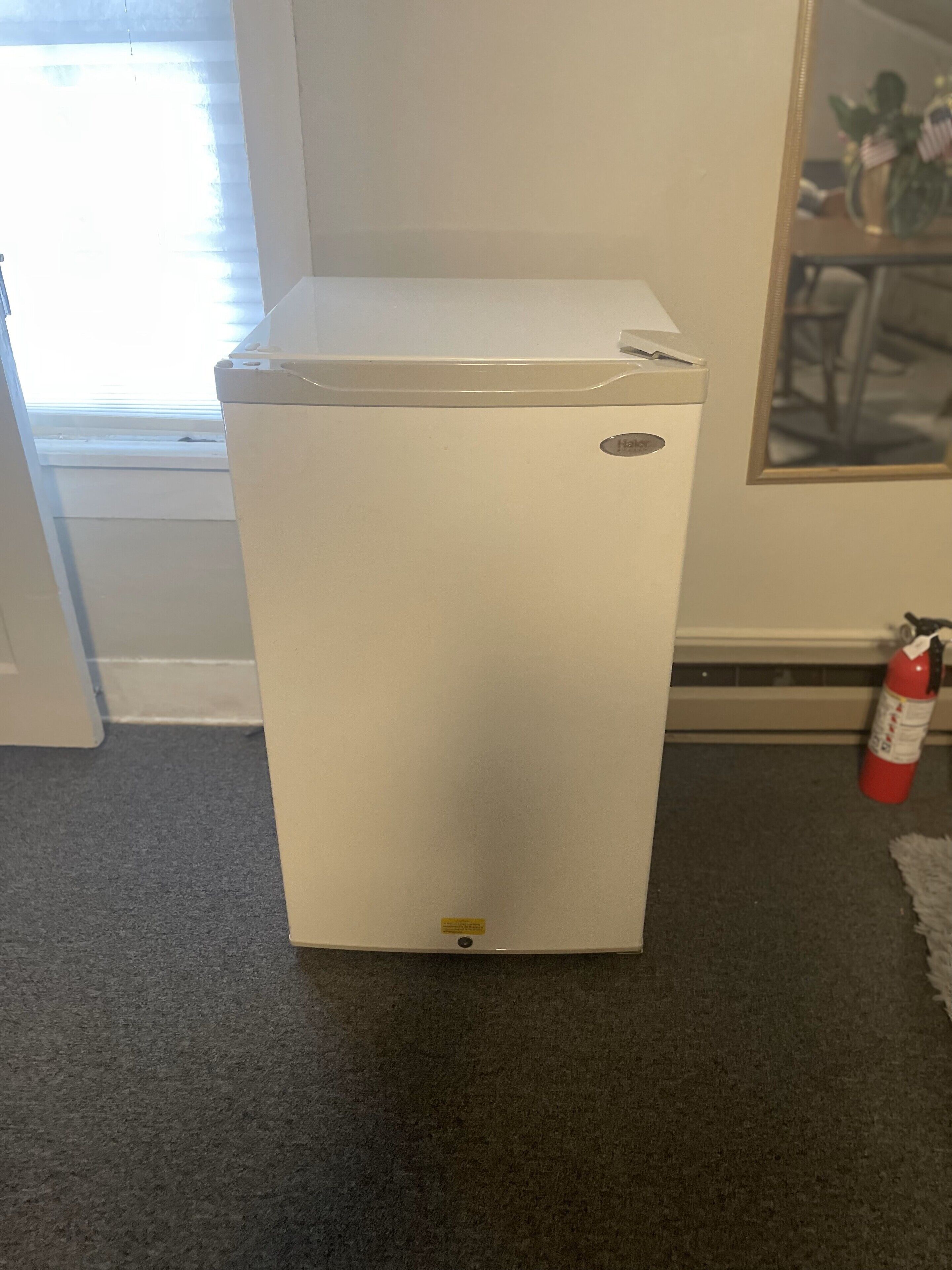 Un refrigerador blanco en una habitación con una ventana, un extintor y un cuadro en la pared.