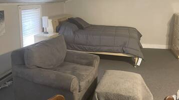 Une petite pièce peu éclairée, avec un lit en mezzanine, un canapé et une chaise en bois.