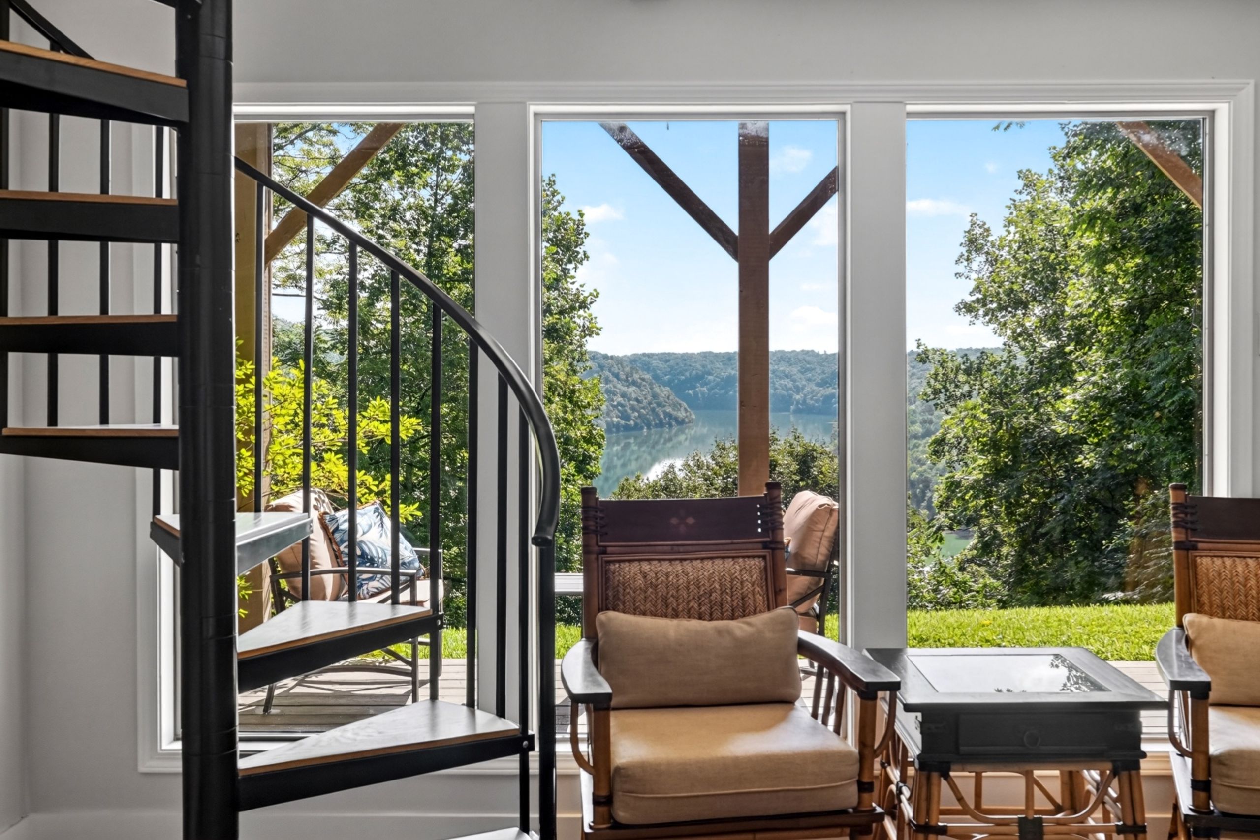 A modern interior with a spiral staircase, a dining area with chairs and a table, and large windows offering a view of a forested landscape.