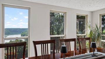 A dining area with a table, chairs, and a view of a river and forest through large windows.