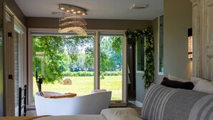 Une chambre à coucher avec un grand lit, une baignoire blanche et une vue sur un jardin verdoyant, visible à travers de grandes fenêtres.