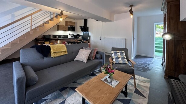 Een moderne woonkamer met een grijze bank, een houten salontafel en een trap die naar een keuken leidt.