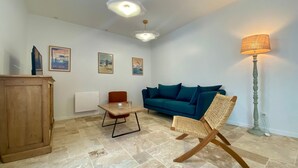A living room with a blue sofa, a wooden cabinet, a wicker chair, and a floor lamp. - Cozy New Cottage Near Beach with Private Terrace, Hot Tub, and EV Charger (Pornic)