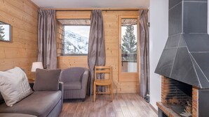 Ein gemütliches Zimmer mit Holzboden, einer Couch, einem Sessel, einem Kamin und einem Fenster mit Vorhängen.
