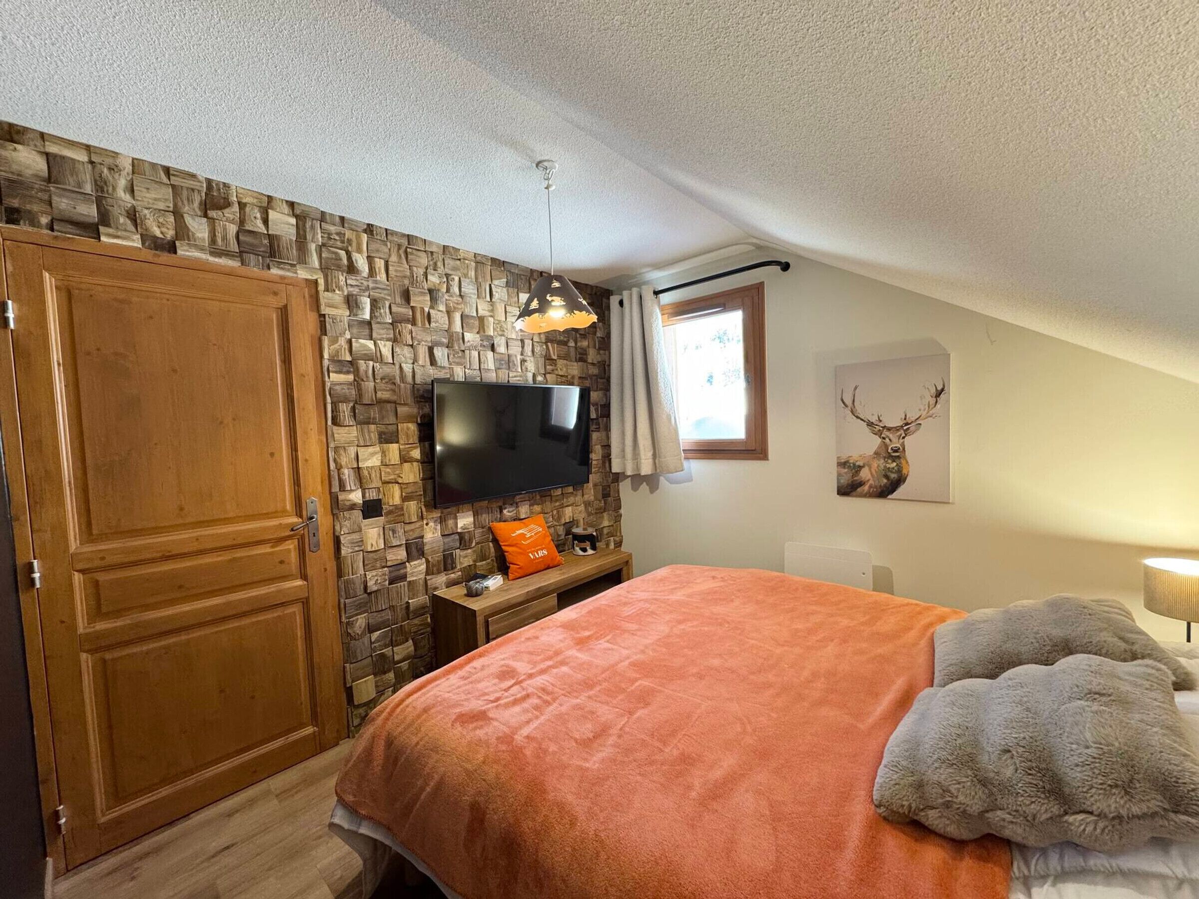 A bedroom with a stone wall, a wooden wardrobe, a bed with an orange blanket, a television, and a deer painting.