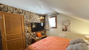 Quarto com parede de pedra, guarda-roupa de madeira, cama com cobertor laranja, televisão e pintura de um veado.