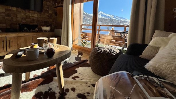 Um quarto aconchegante com parede de madeira, uma janela grande com vista para a montanha, uma mesa redonda com uma garrafa e copos, e um sofá confortável com um travesseiro de pele.