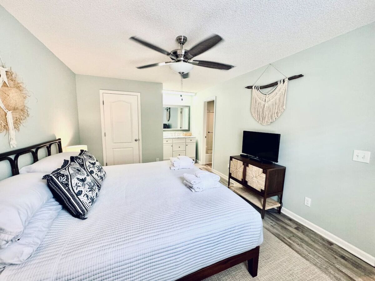 A bedroom with a bed, a ceiling fan, a TV on a wooden stand, and a macrame wall hanging.