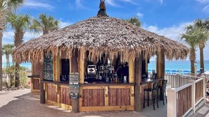 A thatched-roof bar with a wooden counter and chairs, located on a paved walkway with palm trees and a view of the ocean. - Emerald Gem - Gulf Coast View! (Panama City Beach)