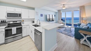 A modern kitchen with white cabinets and stainless steel appliances, a dining area with a blue sofa and a view of the ocean. - Emerald Gem - Gulf Coast View! (Panama City Beach)
