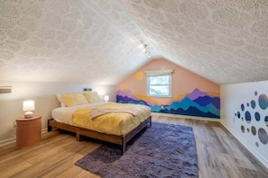 A bedroom with a sloped ceiling, a bed with a yellow bedspread, a nightstand, a window, and a wall mural.