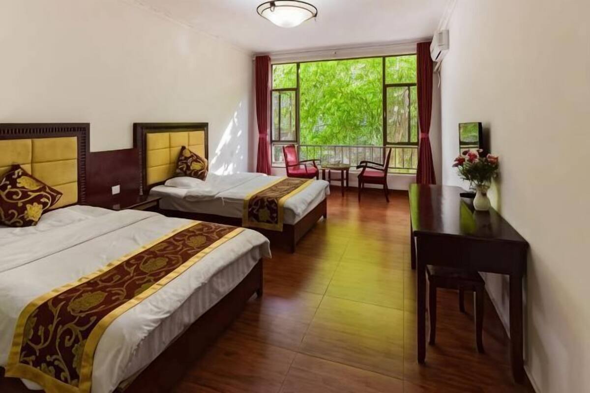 Une chambre d’hôtel avec deux lits, un bureau et une vue sur la verdure grâce à de grandes fenêtres.