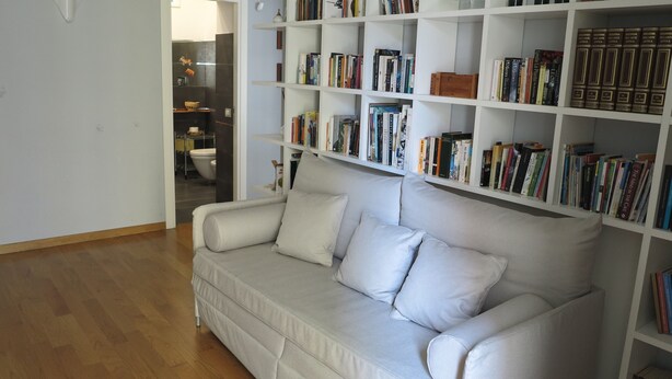 A living room with a white sofa, a bookshelf filled with books, and a wooden floor. - "Nea Parthenope" apartment. (Napoli)