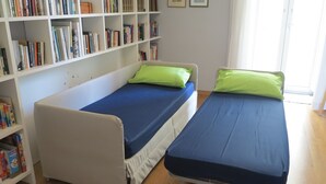 A room with two single beds, each with a blue mattress and a green pillow. There is a bookshelf filled with books to the left of the beds. - "Nea Parthenope" apartment. (Napoli)