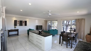 A modern kitchen with white cabinets, a central island, and a dining area with a table and chairs. - Sandcastles Unit 8 - 2 bedroom Ocean Front Unit (Bargara)